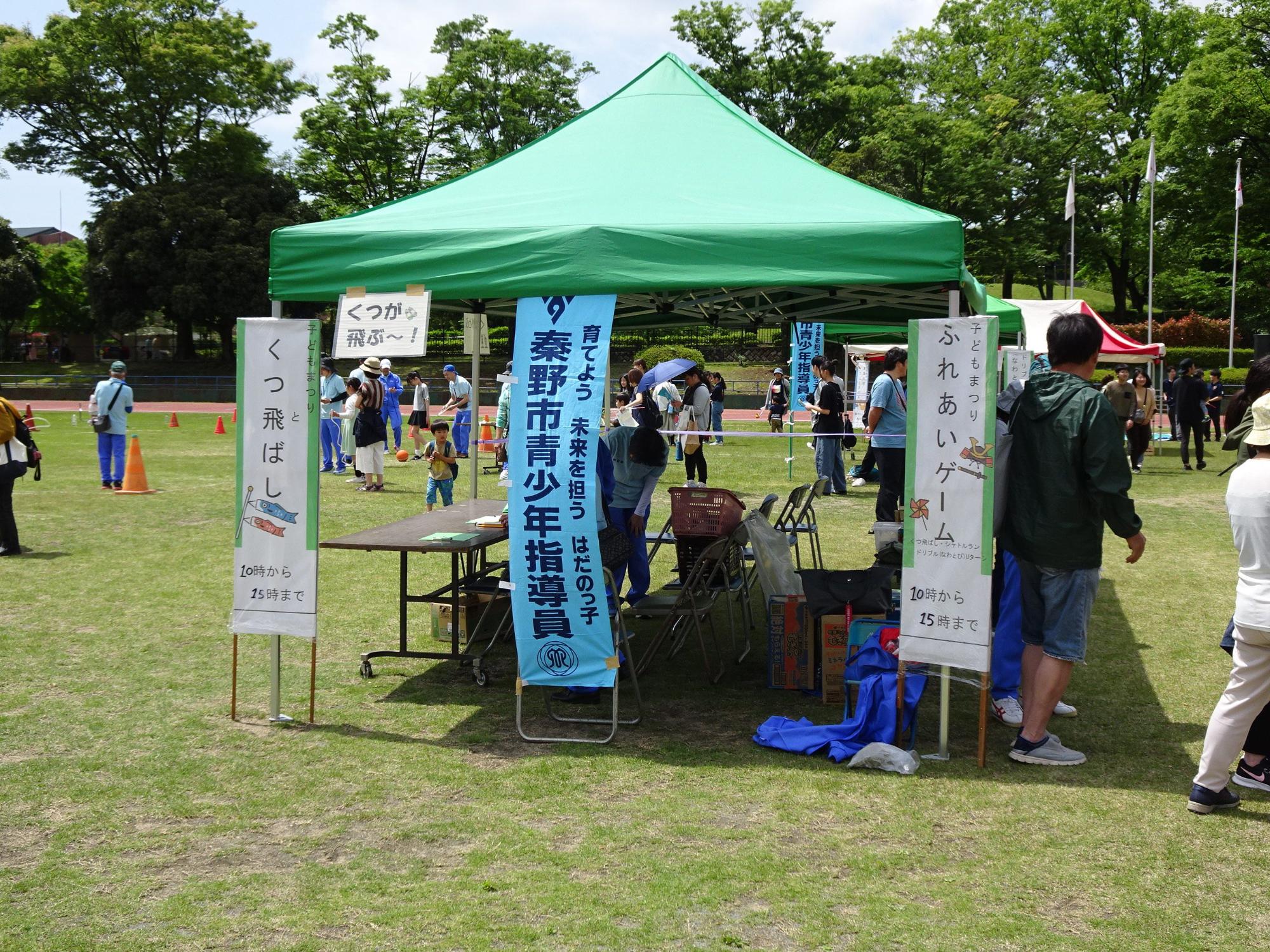 芝生の上の緑色の大きなテントに「秦野市青少年指導員」と書かれたのぼりや「くつ飛ばし」「ふれあいゲーム」の立て看板が設置されており、奥では複数の人達がイベントに参加している様子の写真