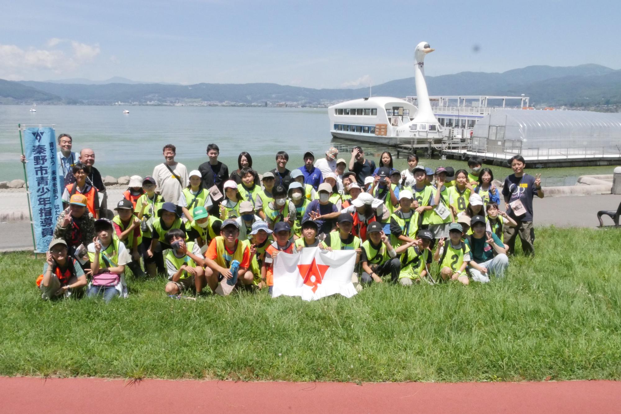 白鳥の遊覧船を背景に、芝生の上で、大人や子供たちがが集合写真を撮っている写真