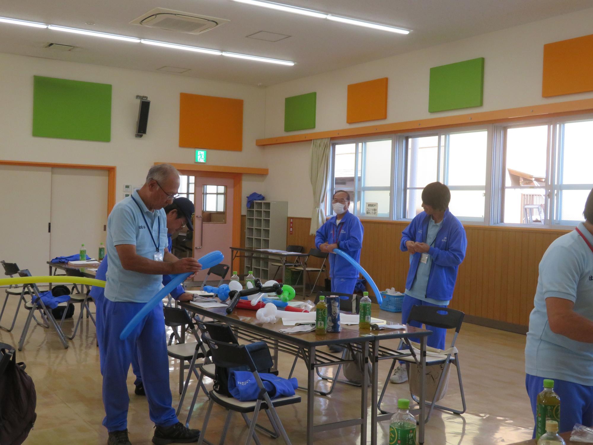 広い室内で複数の人達が、長い風船をねじって犬などの形を作っており、バルーンアートを体験している写真