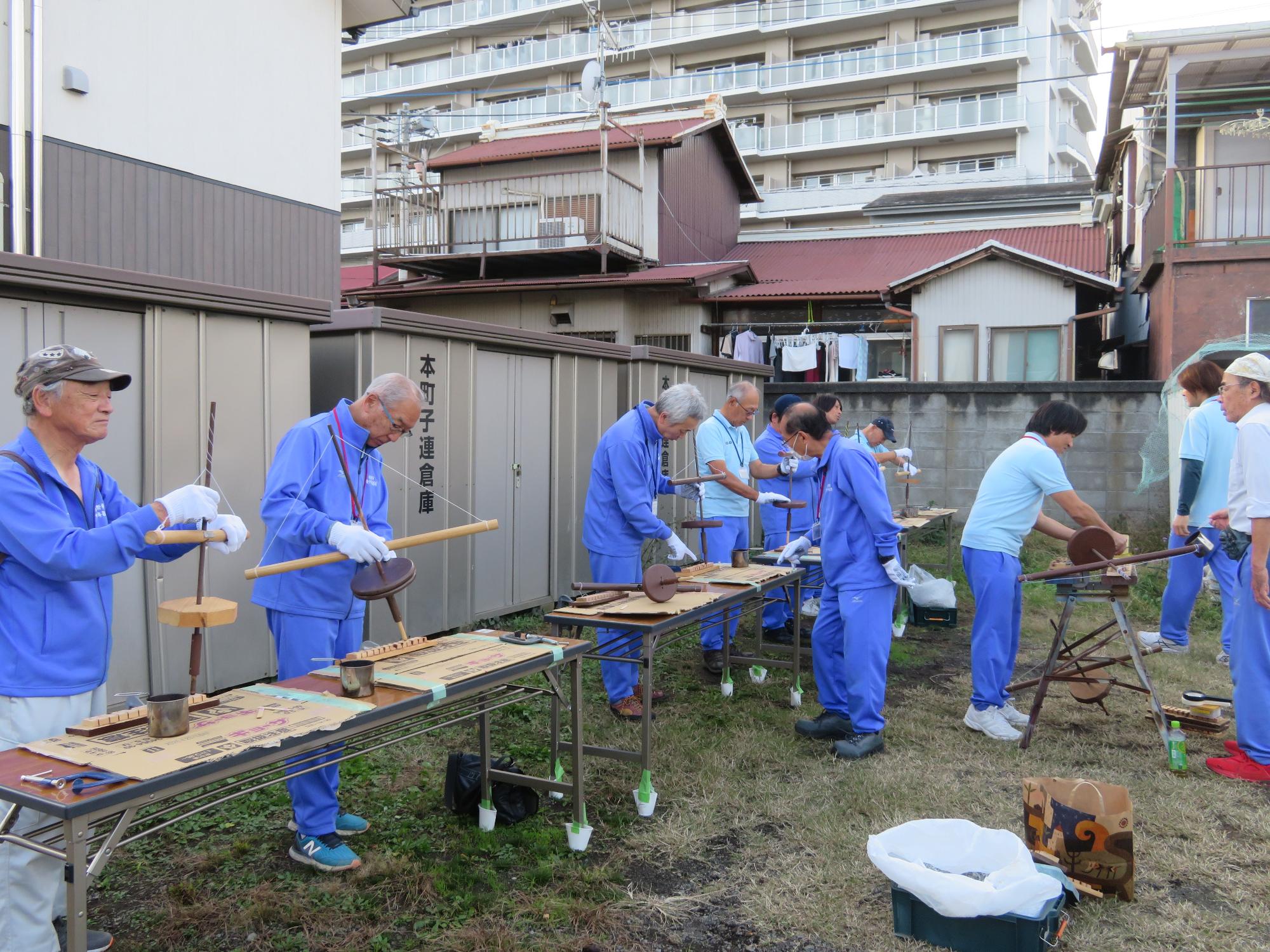 青い作業服を着た複数の人達が、野外で道具を使いながら火起こしを体験している写真