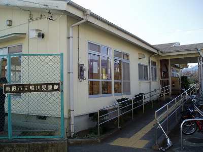 薄い黄色の外壁の平屋建ての建物で、建物の入口横には手すり付きのスロープが設けられており、手前の緑色の金網には「秦野市立堀川児童館」と書かれた看板が取り付けられている堀川児童館外観写真