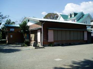 手前の壁に8つの窓が並んでいる茶色の板張りの屋根が白い平屋建ての建物で、左奥には小さな別棟がある堀山下児童館外観写真