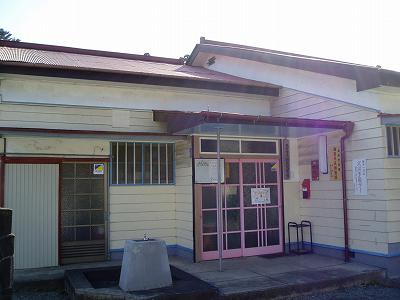 横長のクリーム色をした木造のレトロな平屋建ての建物の正面に赤い屋根の軒下に庇があり、格子状のガラス入りのピンクの引き戸が特徴的な入り口の渋沢児童館外観写真