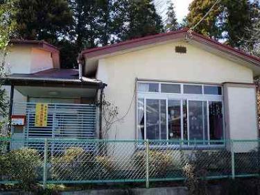 赤い縁取りの屋根を持つ平屋建てのクリーム色の建物で、正面には大きな掃き出し窓と、その左側には緑色の波型フェンスで囲まれた玄関があり、手前には金網フェンスと植栽が配置されている横野児童館外観写真