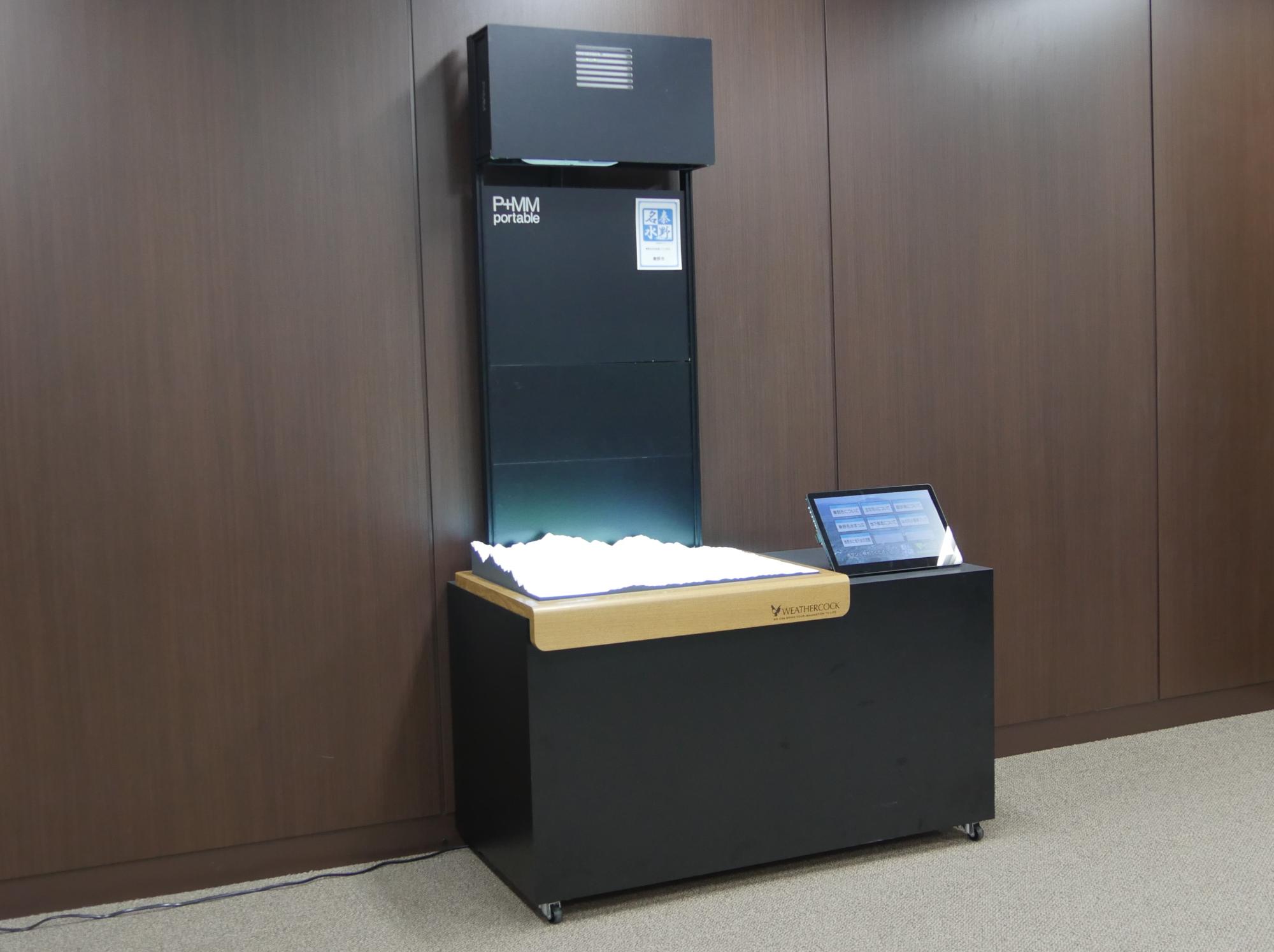 左側に立体地形モデルが置かれ、右側には操作用のタブレットが配置されている黒い筐体の箱状の装置の写真