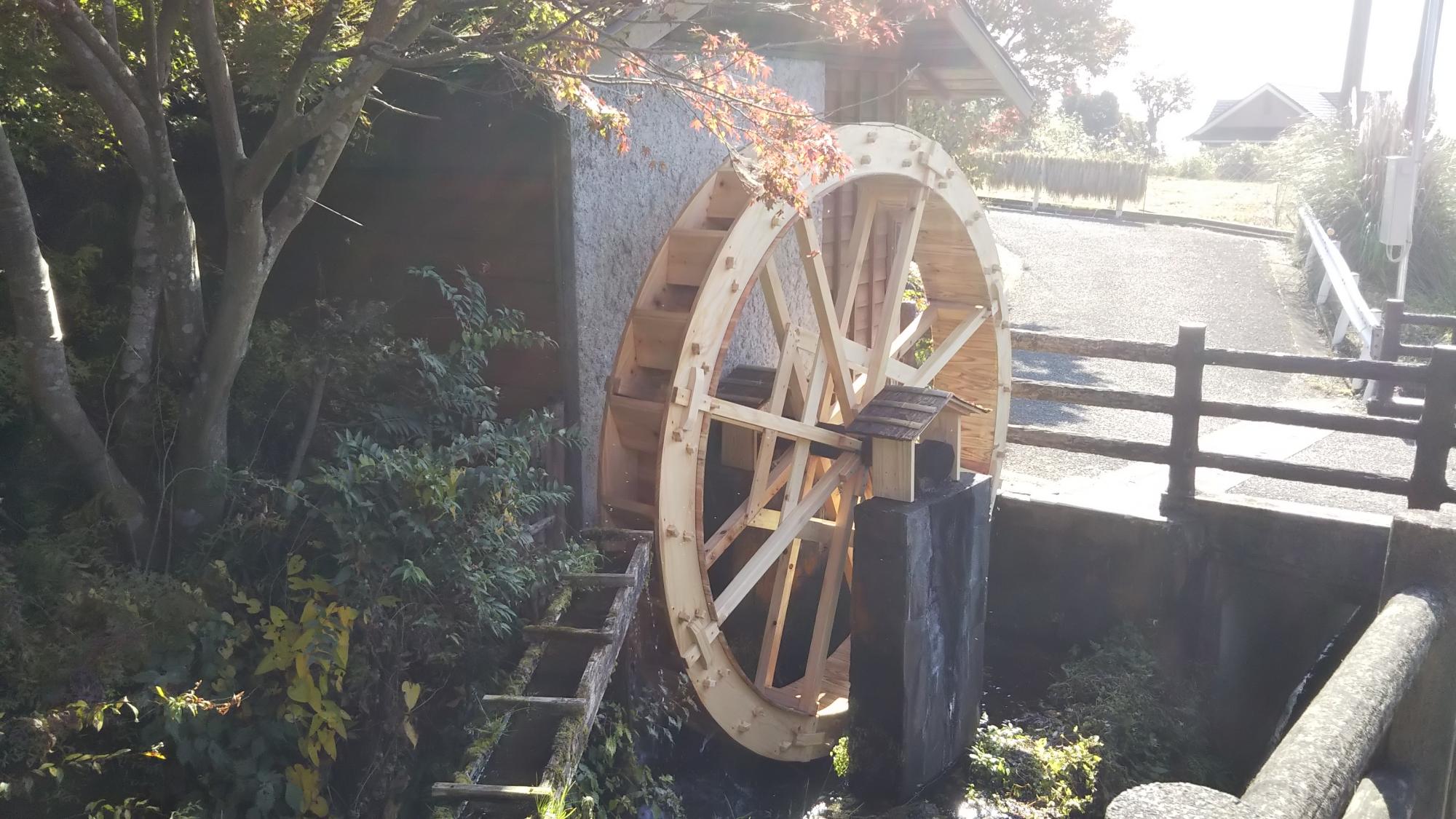 周囲の木々の葉がわずかに紅葉する初秋の強い日差しを浴びて、水車小屋のような建物の壁に半分埋まるように回転している、修繕後の木製の大きな水車の写真