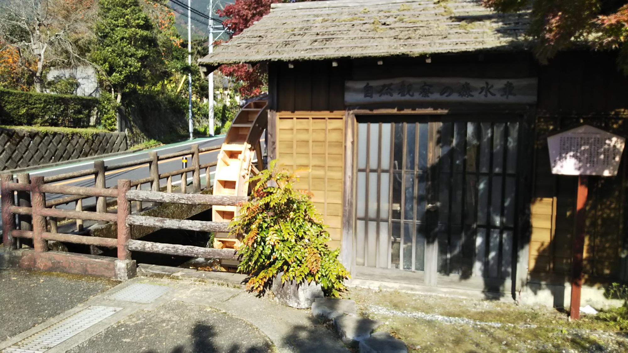 屋根の下に看板が掲げられている渋い木目と茅葺きのような屋根の趣ある小屋の横に小さな木製の水車が設置されている写真
