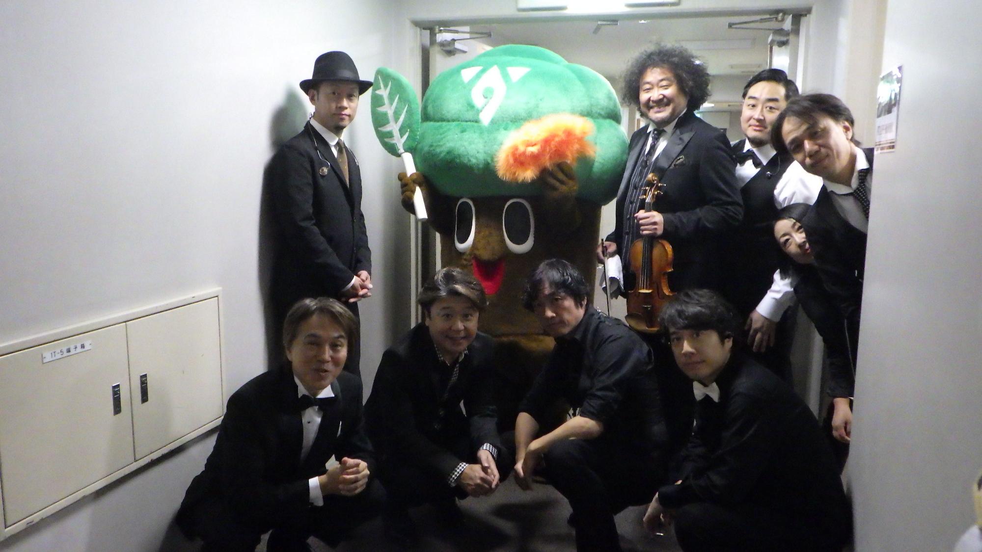 楽屋の前の廊下で、お揃いの黒いスーツを着た8人のメンバーとバイオリンを持った葉加瀬太郎さんが、もりりんと一緒に並んでいる記念写真