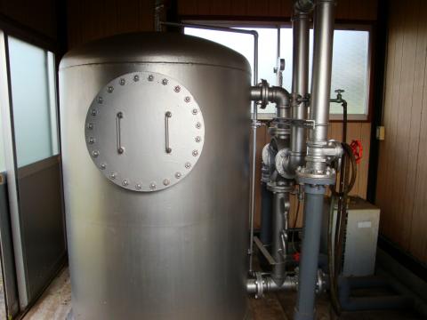 屋内の空間に設置された、銀色の巨大な円筒形のタンクが正面にあり、その右側には複雑に組み合わされた金属製の配管とバルブが見えるろ過機の写真
