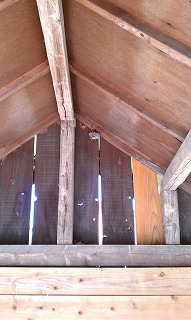 茶色い木材の屋根板と垂木が構成する三角形の空間の、奥の板壁の頂点付近に小さなハチの巣のようなものが一つぶら下がっている写真小屋に作られた蜂の巣