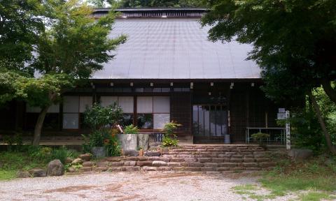 切妻造りのような屋根を持つ、黒っぽい木造の建物の正面を写した写真で、正面中央には石段と玉砂利のような広場があり、両脇と手前には木々や植木が配され、古民家のような静謐な雰囲気を持つ緑水庵の写真