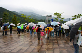 雨が降る曇り空の下、多くの人々が様々な色の傘を差しながら、遠くに山々と吊り橋が見える屋外の石畳の広場に集まっているイベント風景の写真