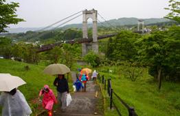 曇りや雨の日に、人々が傘を差しながら草木の生い茂る階段を登っており、遠景には高い主塔と吊り橋が架かった渓谷の景色が広がっている写真
