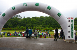 緑の森を背景にした野外会場で、参加者が並んでいる様子を、大きく膨らんだ白いアーチ状の看板越しに撮影した、「第1回春野植樹祭」の開催を知らせるイベント風景の写真