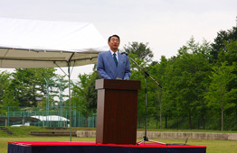 屋外の広大な芝生の会場で、スーツとネクタイを着用した古谷市長が、演台の前に立って、多くの聴衆や木々を背景にして挨拶を行っている、公的なイベント風景の写真