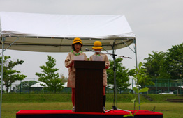 屋外の壇上で、黄色いヘルメットを被った2人の子供が、原稿を読みながら挨拶やメッセージを発表している、式典やイベントでのスピーチ風景の写真