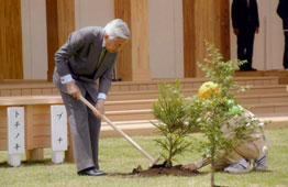 スーツを着た天皇陛下が、鍬を持ってしゃがみ込み、黄色い帽子を被った人物と共に木造建築の前で植樹活動を行っている、公的な行事の一場面を捉えた写真