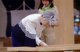 白いトップスを着た皇后陛下が、木製の台に身を乗り出して何かを観察している様子を、背後から作業服姿の若い女性が見守っている、屋内行事の一場面の写真