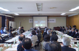 多くの参加者が会議室の机の席につき、正面にあるスクリーンに映し出されたビデオ映像を視聴している会議風景の写真