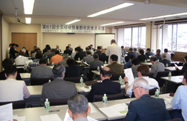 正面には横断幕が掲げられた会議室で参加者の方々が長付けの席につき、資料を広げ会議に参加しており、中央付近に男性が立ち、発言している会議の様子を写した写真