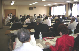 窓から光が入る明るい会議室で、多数の参加者が並べられた長机に着席し、机に広げた資料を見ながら会議に参加している様子を後方から写した写真