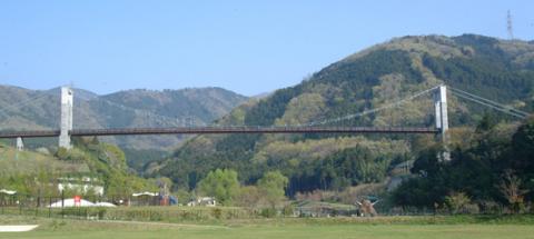 青い空の下、深い山々に囲まれた渓谷に架かる、2つの高いコンクリート製主塔と細長い茶色の歩道を持つ吊り橋が、緑の広場の上に見えている遠景写真