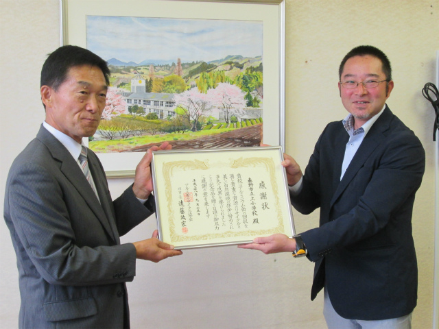 背景に風景画がある部屋の中で、感謝状を二人の男性が持ちカメラに微笑む写真