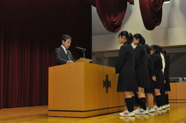 檀上に立つスーツ姿の男性が、紺色の制服を着た6人の女子生徒に向かって表彰状を読み上げている様子を写した写真