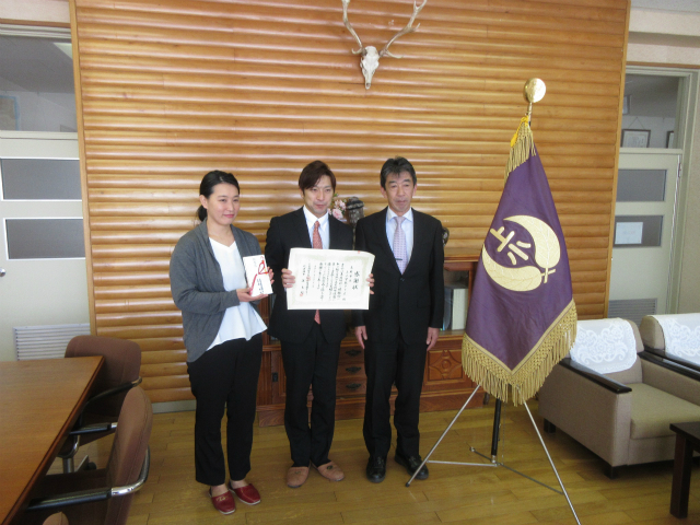 木目調の壁を背景に三人の男女が並んでおり、中央の男性が賞状を持ち、向かって左側の女性は熨斗袋を持っており、右側には校旗が立っている写真