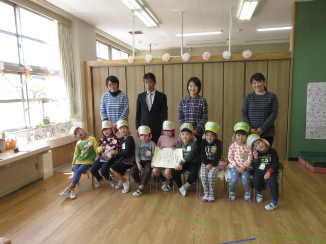 室内で保育士と思われる3人の女性と財団法人の1人の男性、賞状を持った園児を含む9人の園児が、カメラに向かってポーズを取っている集合写真