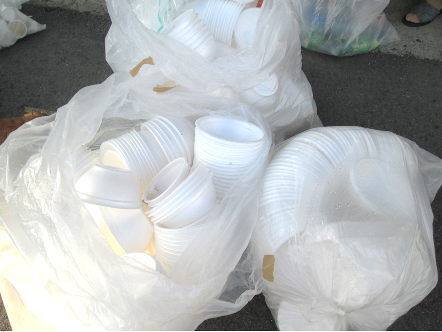 使い終わった白い発泡スチロール製の器やコップが、3つの透明なごみ袋にまとめられて地面に置かれている様子を写した写真