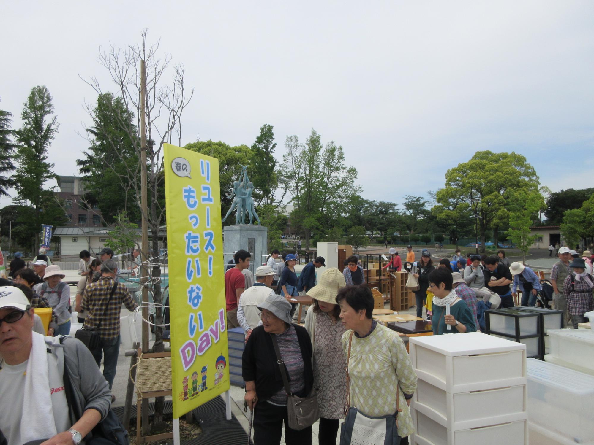 青い空と緑の木々を背景に、リユースイベントの黄色の幟や多くの人々が写っており、賑わっている写真