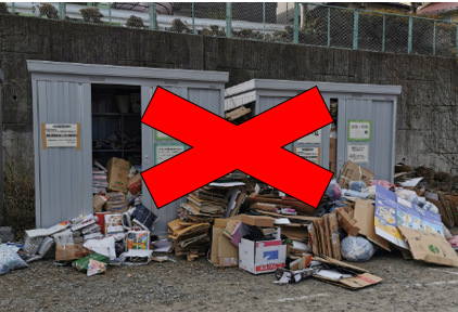 二つの倉庫の引き戸が開き、中から溢れ出た大量の段ボールや古紙が手前に山積みになっており、その全体を覆うように赤い大きなバツ印がつけられた不法投棄されている写真