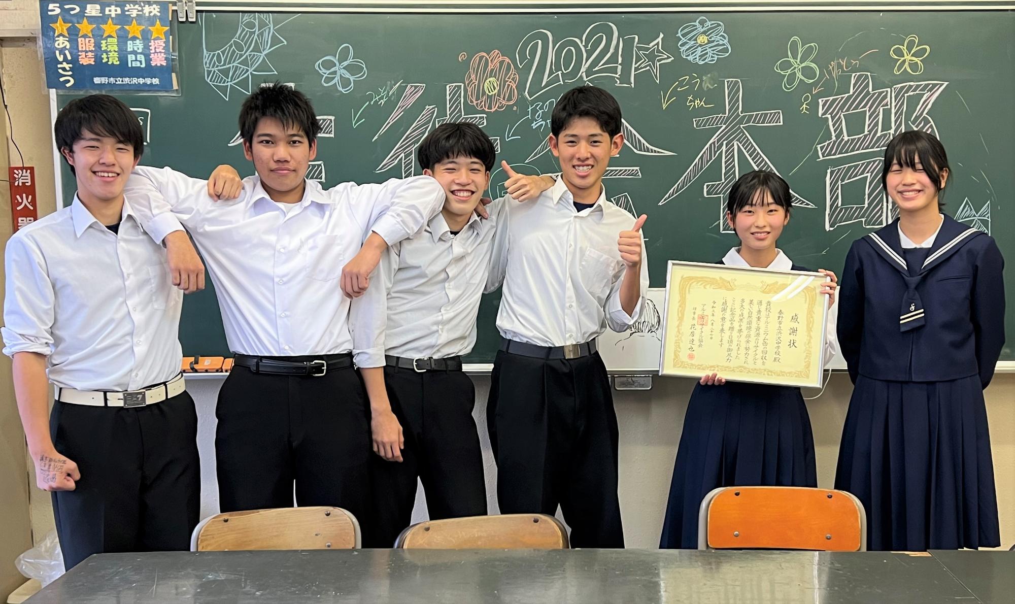 「2021 生徒会本部」と大きく書かれ、花など色とりどりのイラストが描かれた黒板の前で、賞状を持った女子生徒と、笑顔で親指を立てる男子生徒など6人の生徒が並んで写っている記念写真