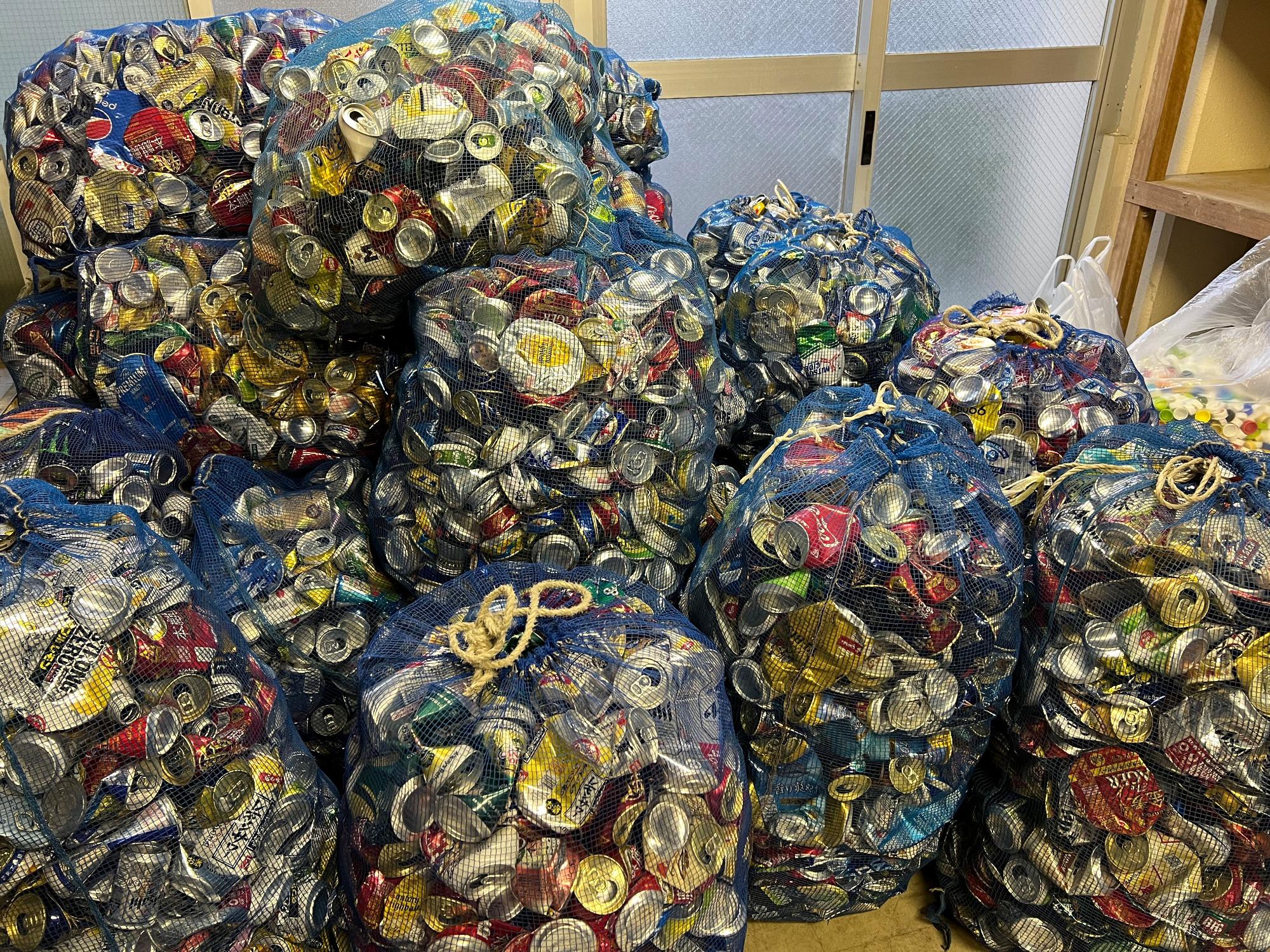 潰された空き缶が網の袋にぎっしりと詰められ、山積みになっている様子を写した写真