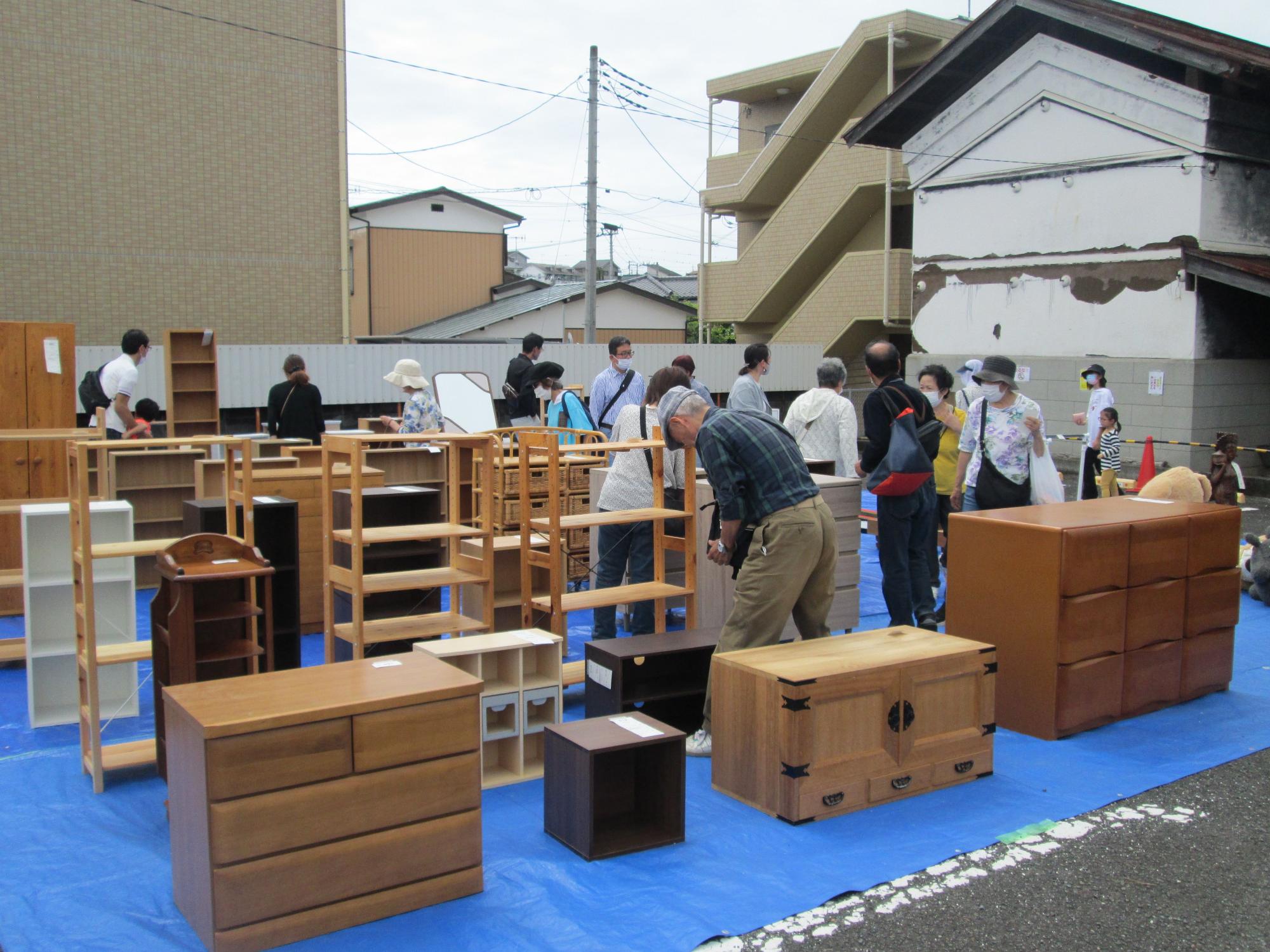 屋外イベント会場の青いビニールシートの上に、整理ダンスや棚など多くの木製家具が並べられ、たくさんの人が品定めをしている様子を写した写真