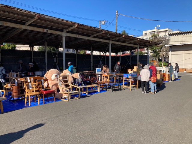 骨組みと屋根だけの建物の下に、木製の椅子やテーブル、大きなクマのぬいぐるみなどのリユース品がブルーシートの上に並べられ、たくさんの人が品定めをしている様子を写した写真