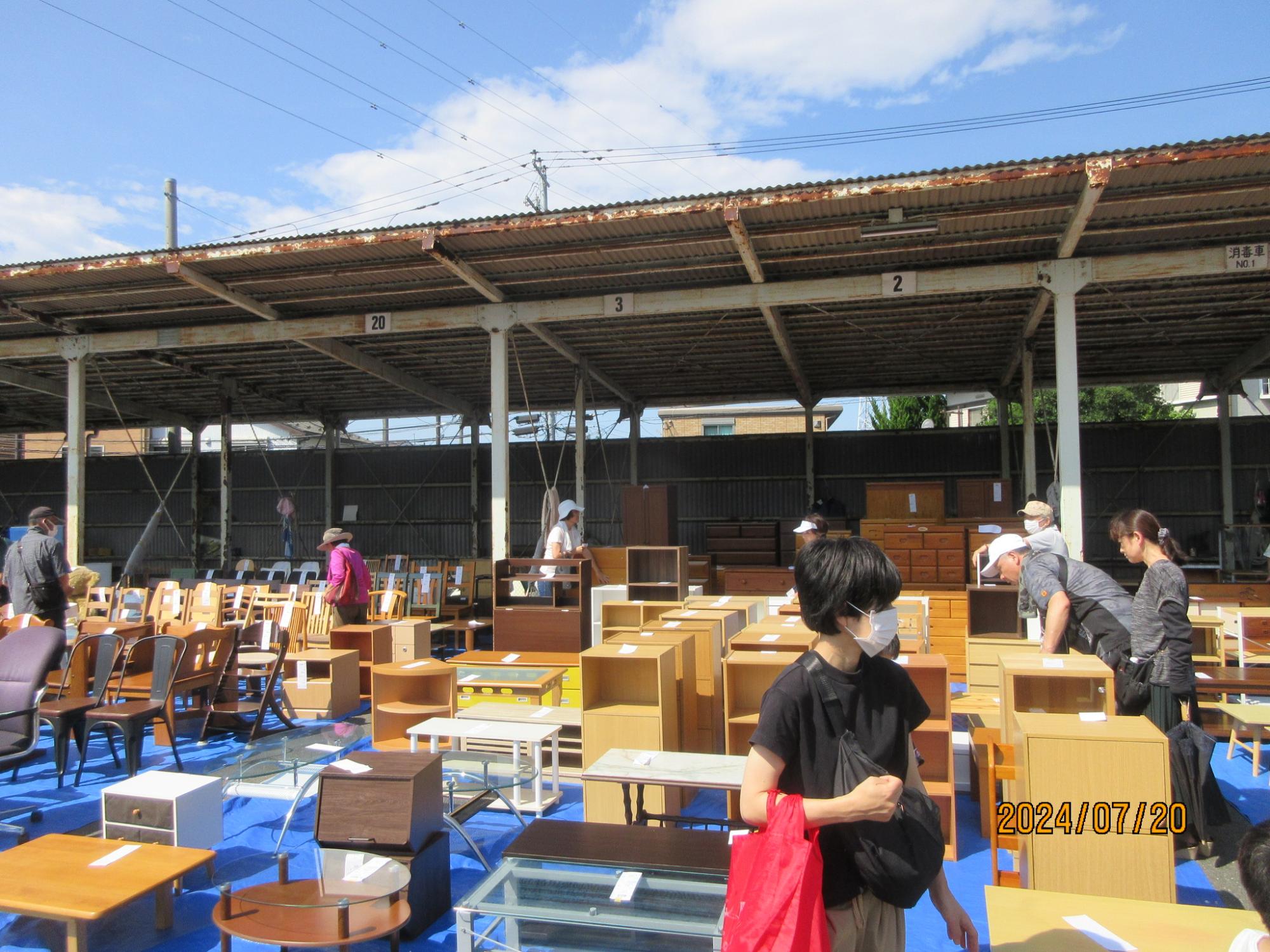 リユース品のイベント会場で、ブルーシートの上に並べられたたくさんの木製の家具や椅子を、多くの人々が品定めをしている様子を写した写真