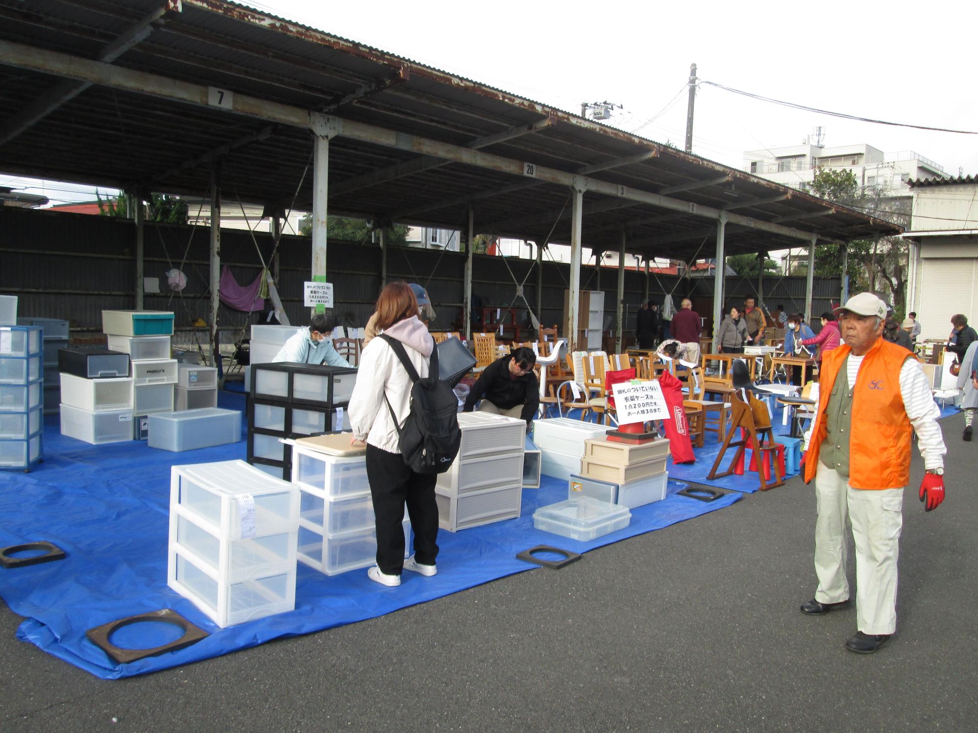 屋外に敷かれた青いビニールシートの上に並べられたリユース品の椅子やプラスチックケースなどを、多くの人々が品定めをしている様子を写した写真