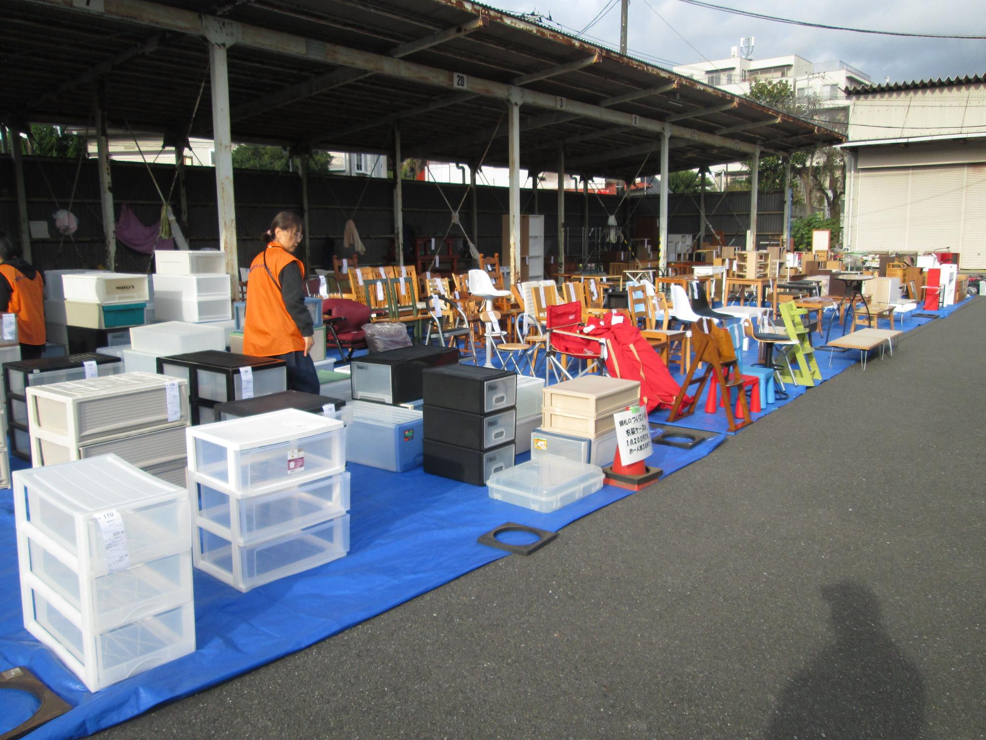 骨組みと屋根だけの建物の下に、プラスチックケースやパイプ椅子などの不用品がブルーシートの上に並べられ、係員が立っている写真