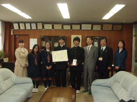 賞状と熨斗袋を持った制服姿の男子生徒2人と女子生徒2人、スーツ姿の大人たち4人が並んで立っている集合写真