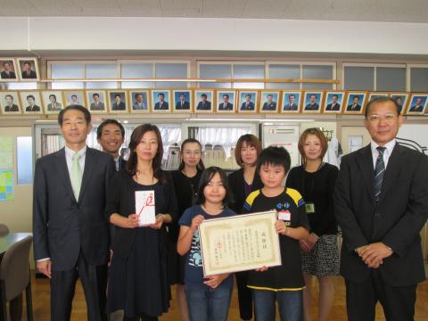 小学校の校長室で、賞状を持った児童二人を中心に、目録の封筒を持った女性、スーツ姿の男性2人が前列に立ち、4人の男女が後列に並んで写っている記念写真