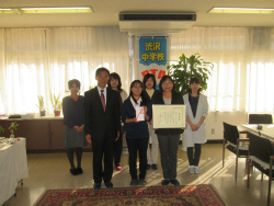 窓から光が差し込む部屋で、前列に賞状と目録の封筒を持った2人の女性とスーツの男性、後列に4人の女性が笑顔で並んでいる集合写真