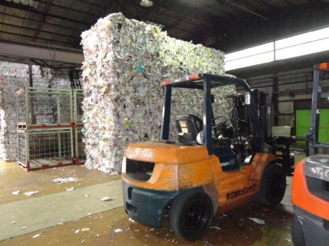 リサイクル工場のような屋内作業場に、天井近くまで積み上げられた巨大な新聞紙や雑紙の圧縮ベールがそびえ立ち、手前にはオレンジ色のフォークリフトが停車している、中間処理の様子の写真