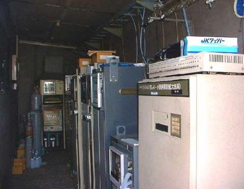 暗い室内に大型の電算機やサーバーラックのような機材が通路を挟んでずらりと並べられており、手前の大きな筐体の上には「JKファイバー」と書かれた青い箱が置かれている機材室の様子の写真