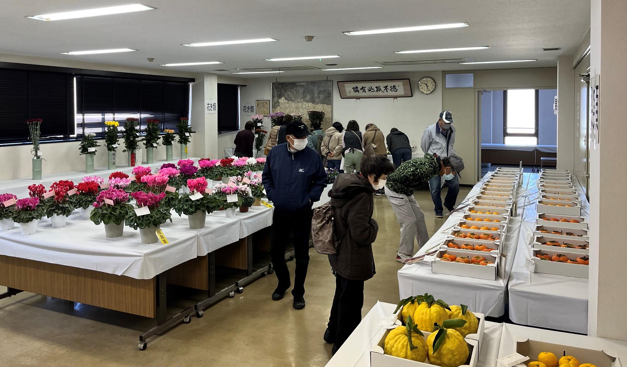 手前のテーブルにはシクラメンやバラの鉢植え、奥のテーブルにはミカンやユズのような果物が陳列されており、マスクを着用した多くの来場者が商品を鑑賞したり購入したりしている様子の写真