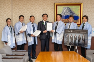 スーツ姿の市長が中央に立ち、その両脇を水色の法被を着た男女6人が取り囲んでおり、要望書やロックバンドLUNA SEAのポスターを持って記念撮影をしている写真