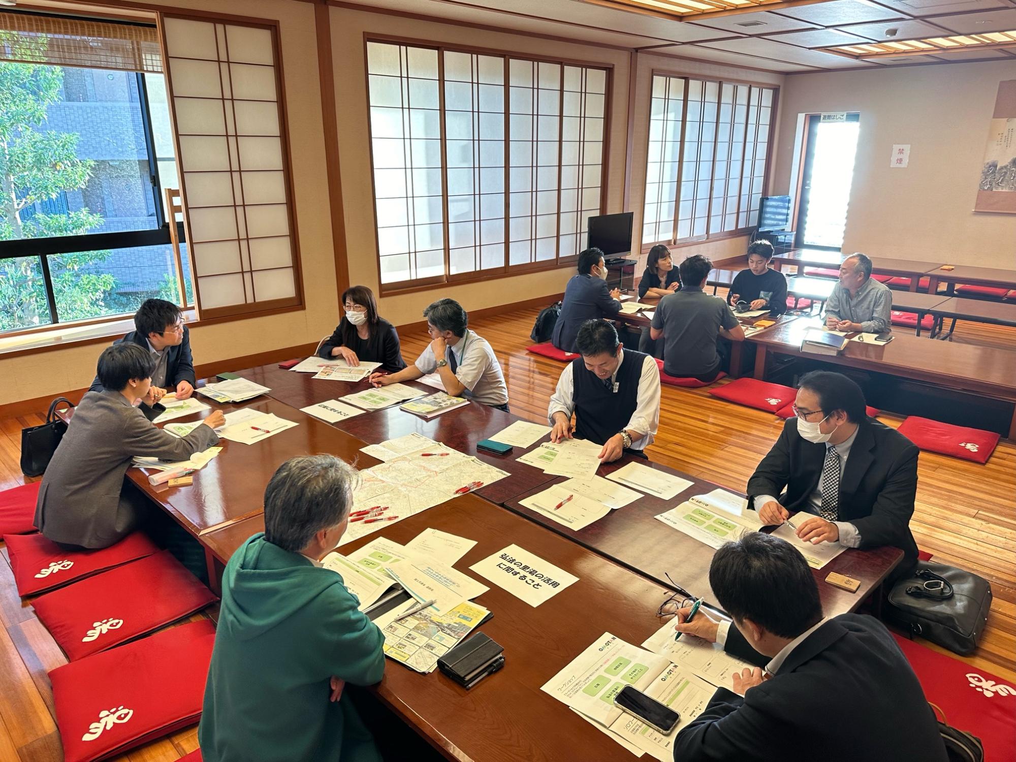 明るく広々とした和風の部屋で、たくさんの参加者たちが大きな木製の座卓を囲んで赤い座布団に座り、資料や地図を広げながら話をしている様子の写真
