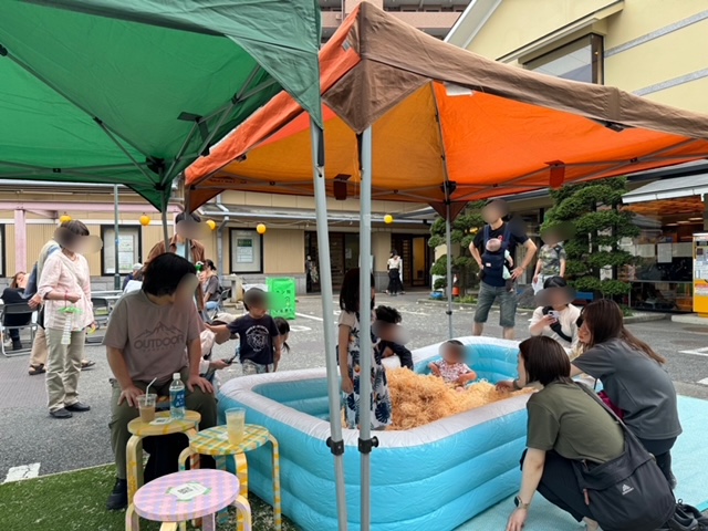 屋外会場の緑とオレンジのテントの下で、木くずが入った水色のビニールプールで数人の子供たちが遊ぶ様子を、保護者やスタッフが周りで見守っている写真