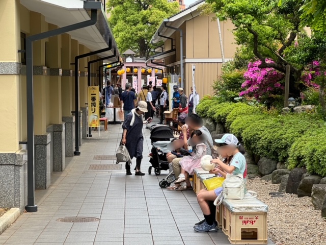 煉瓦色の瓦屋根と柱が並ぶ通路で、手入れされた植栽の前に並んだベンチに複数の来場者が腰掛けている様子の写真